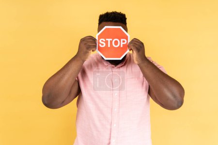Photo for Portrait of man wearing pink shirt covering face with Stop symbol, anonymous person holding red traffic sign, warning about road safety rules. Indoor studio shot isolated on yellow background. - Royalty Free Image