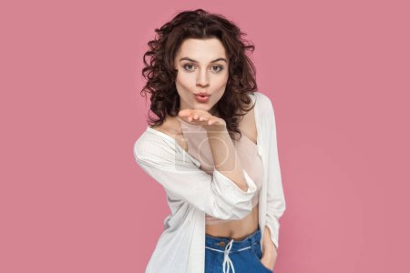 Photo for Portrait of lovely beautiful woman with curly hairstyle wearing casual style outfit sending air kiss over palm, expressing love. Indoor studio shot isolated on pink background. - Royalty Free Image