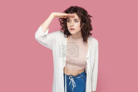 Photo for Portrait of serious attentive woman with curly hairstyle wearing casual style outfit keeps palm over forehead, looking far in future. Indoor studio shot isolated on pink background. - Royalty Free Image