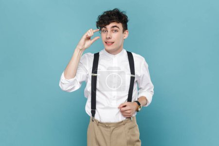 Photo for Portrait of attractive man wearing white shirt and suspender showing little small gesture with fingers, frowning face with disappointed expression. Indoor studio shot isolated on blue background. - Royalty Free Image