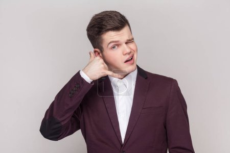 Photo for Portrait of handsome young man makes call me gesture, winking his eyes, flirting with beautiful girl, wearing violet suit and white shirt. Indoor studio shot isolated on grey background. - Royalty Free Image