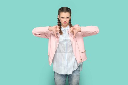 Photo for Portrait of displeased unhappy teenager girl with braids wearing pink jacket standing with thumbs down, showing dislike gesture to camera. Indoor studio shot isolated on green background. - Royalty Free Image