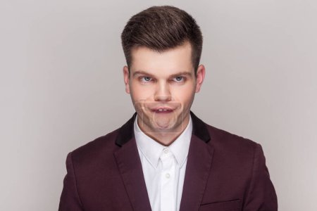 Photo for Portrait of crazy comic foolish handsome young man standing looking at camera with funny crossing eyes, wearing violet suit and white shirt. Indoor studio shot isolated on grey background. - Royalty Free Image