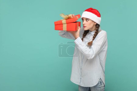 Photo for Portrait of scared teenager girl with braids wearing striped shirt and Santa Claus hat, holding present box, looking inside with big eyes. Indoor studio shot isolated on green background. - Royalty Free Image