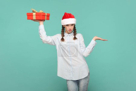 Photo for Portrait of joyful cheerful teenager girl with braids wearing striped shirt and Santa Claus hat, holding present box, presenting copy space on palm. Indoor studio shot isolated on green background. - Royalty Free Image