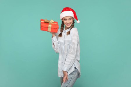 Téléchargez les photos : Portrait d'adolescente heureuse souriante avec des tresses portant une chemise rayée et un chapeau du Père Noël, tenant une boîte-cadeau rouge, regardant la caméra. Studio intérieur tourné isolé sur fond vert. - en image libre de droit