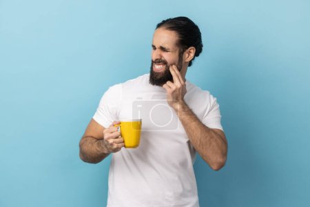 Téléchargez les photos : Portrait d'un homme barbu portant un T-shirt blanc debout et touchant sa joue parce qu'il ressent de la douleur sur la dent, après avoir bu de l'eau froide. Studio intérieur tourné isolé sur fond bleu. - en image libre de droit