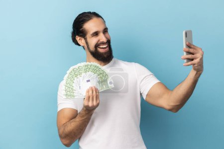 Photo for Portrait of delighted optimistic man with beard wearing white T-shirt taking selfie on cellphone with fan of euro banknotes in hands. Indoor studio shot isolated on blue background. - Royalty Free Image