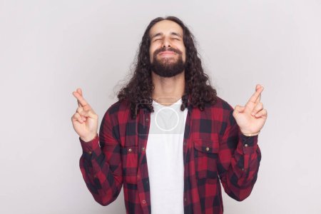 Photo for Glad positive bearded man with long curly hair in checkered red shirt keeps fingers crossed, waits for special moment, closes eyes in anticipation. Indoor studio shot isolated on gray background. - Royalty Free Image