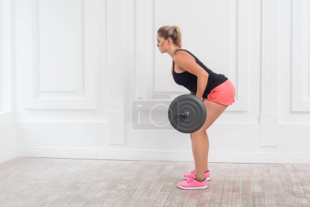 Photo for Side view portrait of athletic strong bodybuilder woman in pink shorts and black top doing exercises for gluteus muscle at gym with the barbell on white wall background. Indoor studio shot. - Royalty Free Image