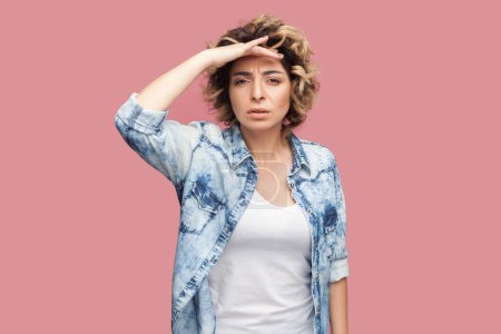 Photo for Portrait of serious attentive woman with curly hairstyle wearing blue shirt standing with palm near forehead, looking, trying to see something. Indoor studio shot isolated on pink background. - Royalty Free Image