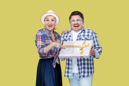 Photo for Happy joyful couple of friends, adult man and woman in casual checkered shirt standing together and holding present box, congratulating. Indoor studio shot isolated, on yellow background - Royalty Free Image