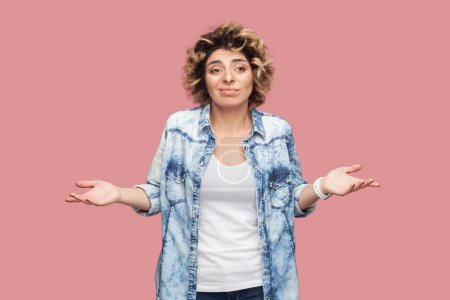 Photo for Portrait of puzzled confused woman with curly hairstyle wearing blue shirt standing shrugging shoulders, having uncertain expression. Indoor studio shot isolated on pink background. - Royalty Free Image