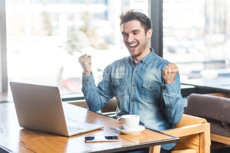 Photo for Portrait of extremely happy positive young man freelancer in blue jeans shirt working on laptop, clenched fists, celebrating his success. Indoor shot near big window, cafe background. - Royalty Free Image