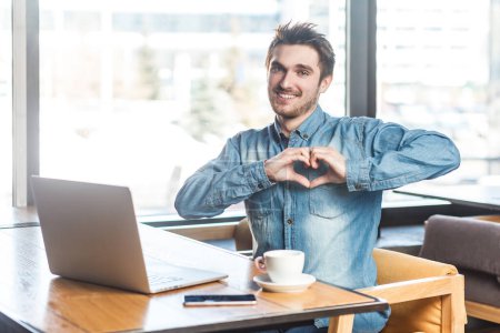 Photo for Portrait of smiling romantic bearded young man freelancer in blue jeans shirt working on laptop, showing heart shape, love gesture to camera. Indoor shot near big window, cafe background. - Royalty Free Image