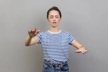 Photo for I'm blind. Portrait of woman wearing striped T-shirt walking with closed eyes, stretching hands to search way in darkness, vision problems. Indoor studio shot isolated on gray background. - Royalty Free Image