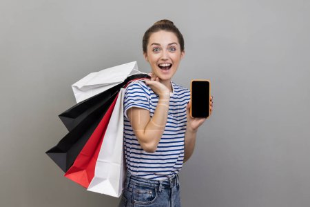 Photo for Portrait of amazed woman holding shopping bags and cell phone, advertising application for online ordering, purchase and delivery of goods. Indoor studio shot isolated on gray background. - Royalty Free Image