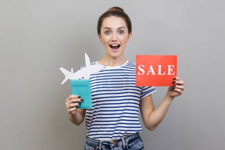 Photo for Portrait of excited woman wearing striped T-shirt holding passport with paper air plane and sale word sign, looking at camera with amazement, traveling. Indoor studio shot isolated on gray background. - Royalty Free Image