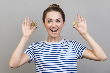 Photo for Portrait of excited woman wearing striped T-shirt standing holding bitcoins, paying attention at new digital cryptocurrency. Indoor studio shot isolated on gray background. - Royalty Free Image