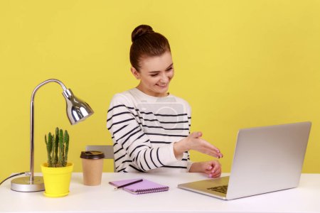 Photo for Satisfied woman office manager welcoming client with handshake looking at laptop screen, talking on video call, online conference. Indoor studio studio shot isolated on yellow background. - Royalty Free Image
