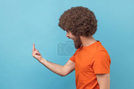Foto de Vista lateral del hombre enojado con peinado afro en camiseta naranja mostrando el dedo medio y pidiendo que se baje expresando negatividad, comportamiento irrespetuoso. Estudio interior plano aislado sobre fondo azul. - Imagen libre de derechos