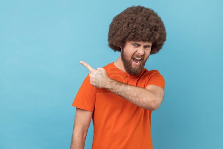 Photo for Portrait of man with Afro hairstyle wearing orange T-shirt pointing to the side and turning away with angry vexed face, giving order to leave. Indoor studio shot isolated on blue background. - Royalty Free Image