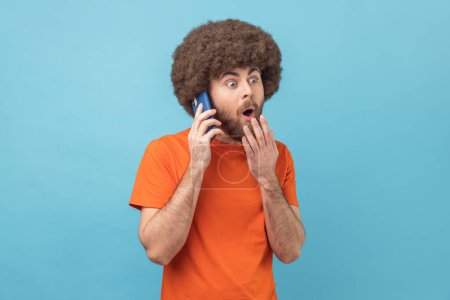 Photo for Portrait of shocked man with Afro hairstyle wearing orange T-shirt talking phone, calling to discuss gossips, affordable tariffs, roaming. Indoor studio shot isolated on blue background. - Royalty Free Image