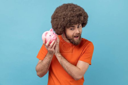 Photo for Portrait of curious excited man with Afro hairstyle wearing orange T-shirt holding shaking piggy bank, being interested how much money inside. Indoor studio shot isolated on blue background. - Royalty Free Image
