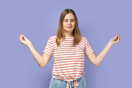 Photo for Portrait of peaceful blond woman wearing striped T-shirt relaxing and doing meditation gesture with fingers, practicing yoga. Indoor studio shot isolated on purple background. - Royalty Free Image