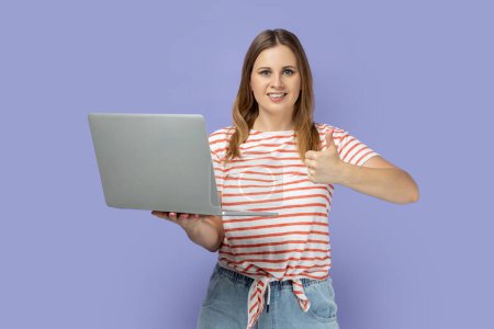 Téléchargez les photos : Portrait de joyeuse femme blonde heureuse portant un T-shirt rayé travaillant sur un ordinateur portable, regardant la caméra et montrant le pouce levé, comme son travail. Studio intérieur tourné isolé sur fond violet. - en image libre de droit