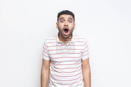 Photo for Portrait of shocked surprised bearded man wearing striped t-shirt standing looking at camera with open mouth and big eyes, sees something astonished. Indoor studio shot isolated on gray background. - Royalty Free Image