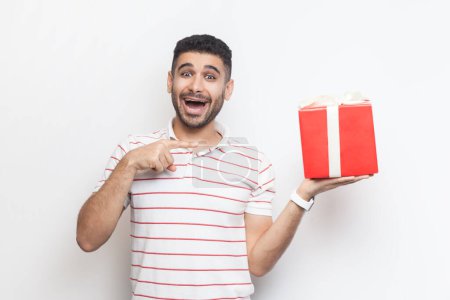 Téléchargez les photos : Portrait d'un homme barbu surpris et excité portant un t-shirt rayé tenant une boîte cadeau enveloppée de rouge, pointant du doigt le cadeau. Studio intérieur tourné isolé sur fond gris. - en image libre de droit