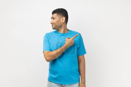 Photo for Portrait of sad upset depressed unshaven man wearing blue T- shirt standing pointing finger away, saying go away, turning his face from camera. Indoor studio shot isolated on gray background. - Royalty Free Image