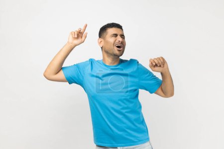 Photo for Portrait of happy excited optimistic unshaven man wearing blue T- shirt standing with raised arms, dancing and singing, celebrating holiday. Indoor studio shot isolated on gray background. - Royalty Free Image