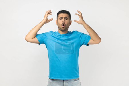 Photo for Portrait of shocked tired exhausted unshaven man wearing blue T- shirt standing showing mind blowing gesture, having annoyed and nervous expression. Indoor studio shot isolated on gray background. - Royalty Free Image