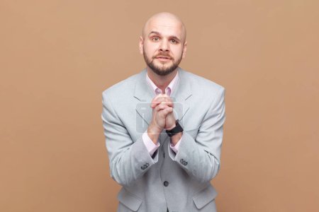 Photo for Hopeful sad uneasy bald man begs for forgiveness, keeps palms in praying gesture, asks mercy and says please, needs your help, wearing gray jacket. Indoor studio shot isolated on brown background. - Royalty Free Image