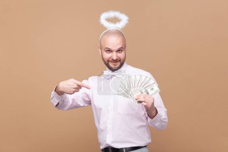 Photo for Portrait of rich bearded man with nimb over head, holding and pointing at dollar banknotes, looking at camera, wearing light pink shirt and bow tie. Indoor studio shot isolated on brown background. - Royalty Free Image