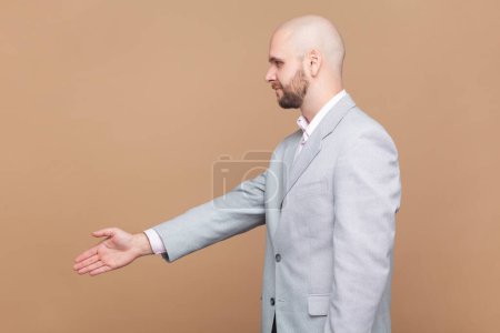 Photo for Side view portrait of bald bearded man reaching out hand to handshake, getting acquainted at job interview, meeting new people, wearing gray jacket. Indoor studio shot isolated on brown background. - Royalty Free Image