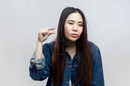 Photo for Portrait of brunette woman in denim jacket standing demonstrates something very little, shapes with fingers small object, feels joyful and optimistic. Indoor studio shot isolated on gray background. - Royalty Free Image
