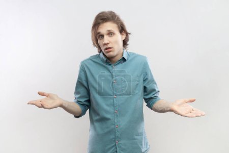 Photo for Portrait of uncertain puzzled confused attractive young man standing spread arms, shrugging shoulders, doesn't know answer, wearing blue shirt. Indoor studio shot isolated on gray background. - Royalty Free Image
