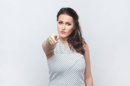 Photo for You are guilty in my misfortune. Portrait of beautiful woman blames someone, points directly at camera, makes serious face, wearing striped dress. Indoor studio shot isolated on gray background. - Royalty Free Image