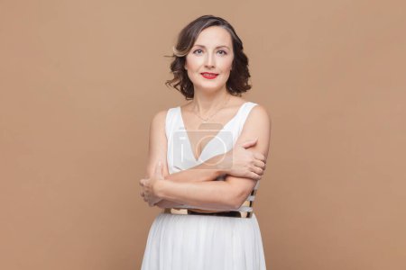 Photo for Portrait of smiling unhappy woman with wavy hair hugging herself, looking at camera, dreaming about something pleasant, wearing white dress. Indoor studio shot isolated on light brown background. - Royalty Free Image