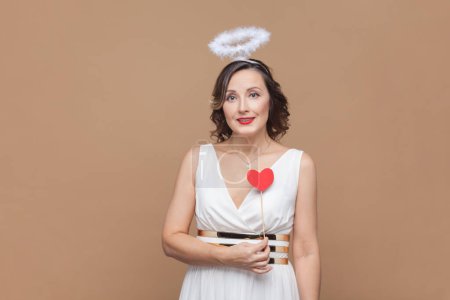 Photo for Portrait of romantic angelic woman with wavy hair and nimb over head, holding little red heart on stick, looking at camera, wearing white dress. Indoor studio shot isolated on light brown background. - Royalty Free Image