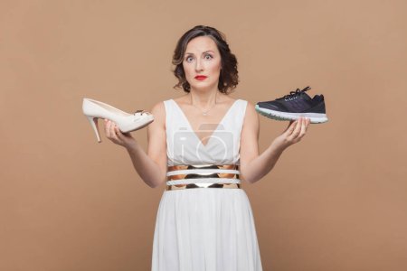 Photo for Woman with wavy hair holding high heels and sneakers and looking with shocked face to camera, dosen't want to take on, wearing white dress. Indoor studio shot isolated on light brown background. - Royalty Free Image
