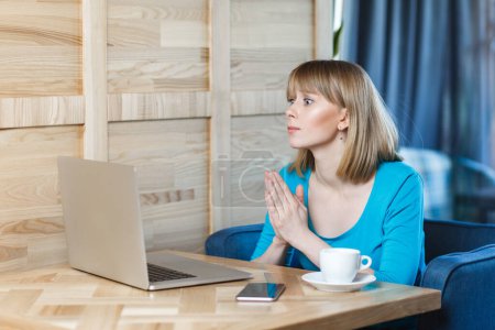 Photo for Side view of hopeful woman with blonde hair in blue shirt working on laptop, having online conversation with boss, asking to continue deadline. Indoor shot in cafe with big window on background. - Royalty Free Image
