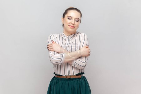 Photo for Portrait of pleased pretty positive woman wearing striped shirt and green skirt standing hugging herself with closed eyes, expressing love. Indoor studio shot isolated on gray background. - Royalty Free Image