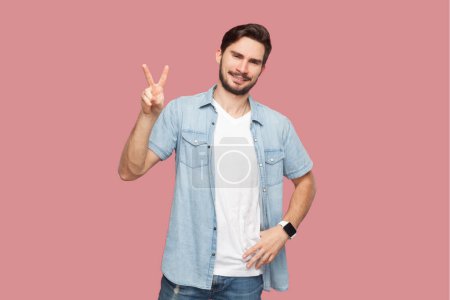 Photo for Portrait of joyful cheerful bearded handsome man in blue casual style shirt standing makes v sign shows peace gesture has glad expression. Indoor studio shot isolated on pink background. - Royalty Free Image