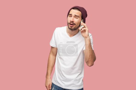 Photo for Portrait of funny cool bearded man in white T-shirt and beany hat standing holding dollar banknotes near ear, pretends talking phone. Indoor studio shot isolated on pink background. - Royalty Free Image
