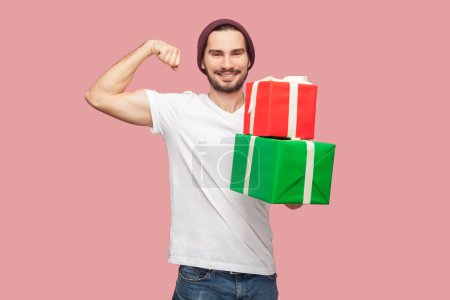 Photo for Portrait of smiling optimistic bearded man in white T-shirt and beany hat standing looking at camera, holding present boxes, showing biceps, raised arms. Indoor studio shot isolated on pink background - Royalty Free Image