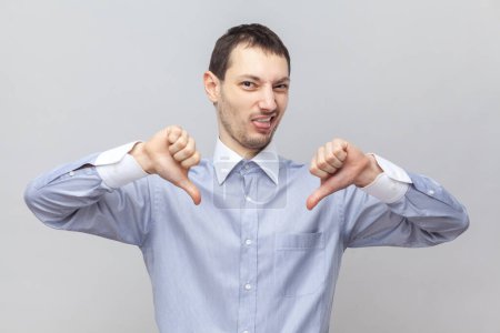 Photo for Portrait of disgusted disappointed handsome man standing showing thumbs down, demonstrates dislike gesture, wearing light blue shirt. Indoor studio shot isolated on gray background. - Royalty Free Image